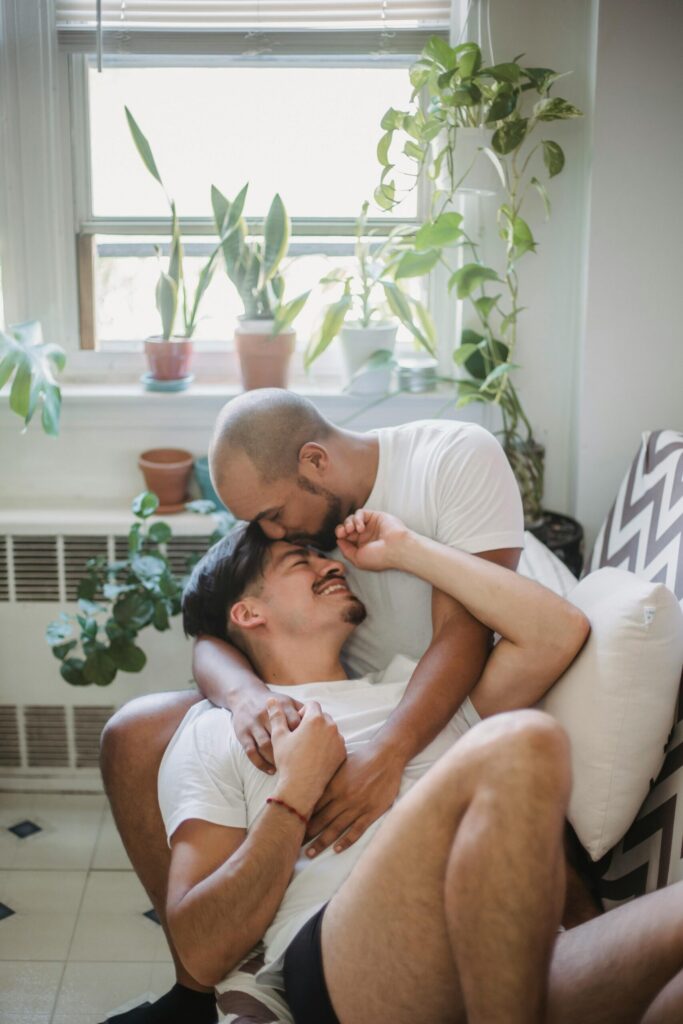 A loving interracial gay couple sharing an affectionate embrace with house plants by the window.
